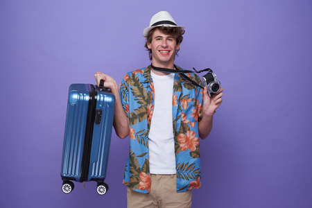 young man tourist with luggage and camera isolated on purple studio background. Tourist having cheerful holiday trip.の写真素材