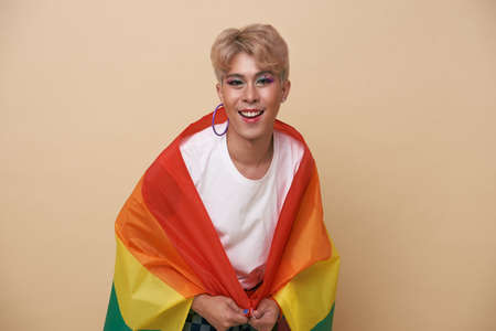 Youth asian transgender LGBT with Rainbow flag on shoulder isolated over nude color background....
