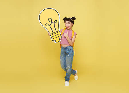 Happy Asian child student holding light bulb with schoolbag isolated on yellow background. idea...