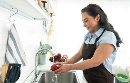 Happy smiling asian older woman in apron washing the apple in kitchen. housewife cooking concept.
