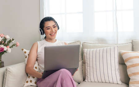 smiling young asian businesswoman using laptop computer while relaxing on a couch at home. work from...