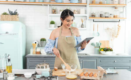 young Asian housewife in kitchen using tablet for searching recipes online cooking the bakery dough...