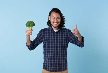 young cheerful asian bearded man showing broccoli and hand thumbs up isolated on blue background.の写真素材