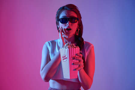 Young woman wearing 3D glasses holding popcorn and beverage watching exciting movie isolated on neon background.の写真素材
