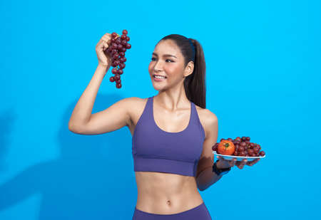 Happy asian woman in sportswear eating fresh fruit gain energy for workout on blue background. Sport, wellbeing and healthy food lifestyle concept.の写真素材