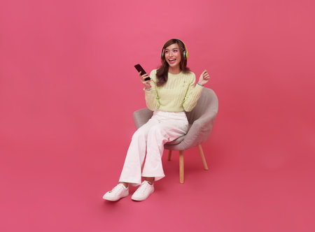 Happy young Asian teen woman smiling listening music in headphones and sitting on chair isolate on pink background.の写真素材