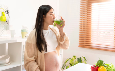 Young Asian pregnant woman drinking green vegetable juice in kitchen room. eating to maintain good health during pregnancy until near childbirth.の写真素材