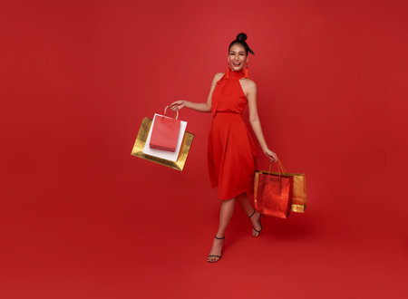 Happy Asian shopaholic woman wearing red dress holding shopping bag walk shopping center isolated on red background.の写真素材