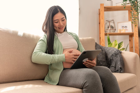Young asian pregnant woman hands fondle on belly and holding tablet sitting on sofa at home. doctor...