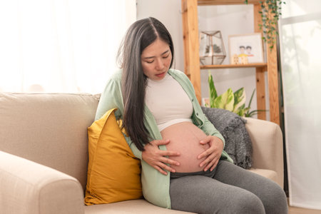Asian young pregnant woman suffering belly ache sitting on a couch in the living room at home.