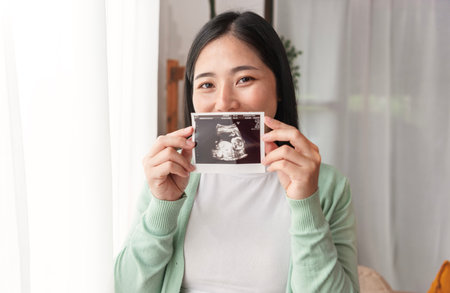 Asian pregnant smiling woman hand showing ultrasound scans at home. Pregnancy, maternity,...