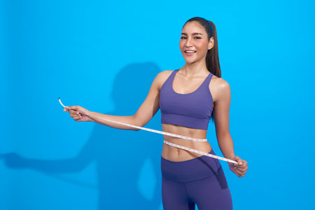 happy smiling asian fitness woman with slim body measuring her waistline and torso isolated on blue studio background. Healthy nutrition and weight losing concept.の写真素材