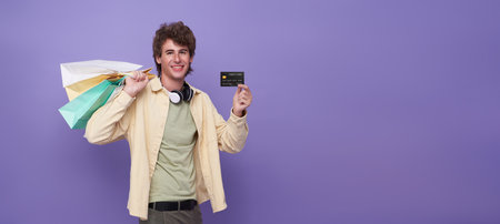 happy smiling handsome man carrying shopping bags and showing credit card in light purple isolated copy space studio background.の写真素材