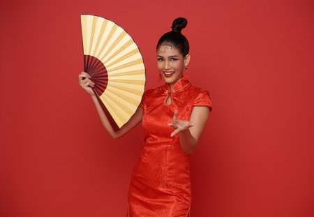 Happy Chinese new year. Beautiful Asian woman wearing traditional cheongsam qipao dress  head holding golden fan in isolated studio red backgroundの写真素材