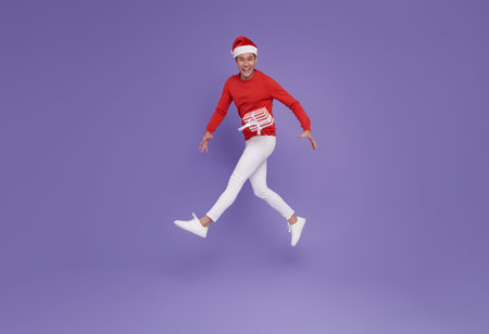 Happy Asian man in santa hat jumping in air with present box in his hands isolated on purple background. christmas celebrationの写真素材