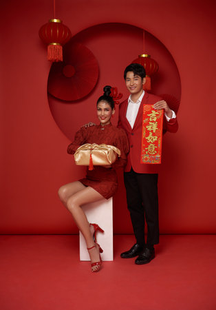 Happy asian couple in red casual attire holding gift box and spring festival couplet bless for Chinese new year day on red room decoration background. Text means great luckの写真素材
