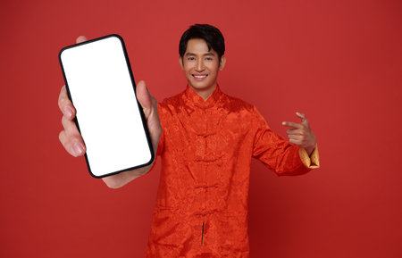 Happy Asian man wearing traditional dress showing mobile phone blank screen isolated on red background.の写真素材
