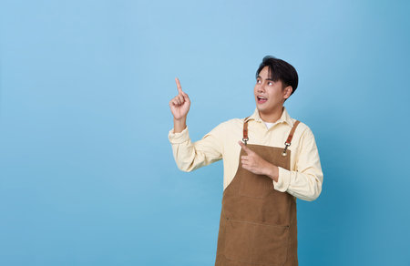 Portrait young asian barista man wearing apron standing and pointing finger advertising isolated on blue background, waitress or entrepreneur cheerful, small business or startup,の写真素材
