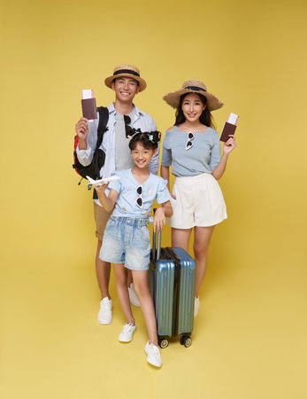 Happy fun asian family vacation portrait. Father, mother and daughters ready for travel flight with suitcase isolated on yellow studio background.の写真素材