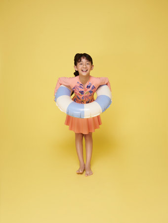 Little funny happy asian girl in swimsuit jumping up in air with inflatable ring around waist, isolated on yellow studio background. Summertime, vacation an school holidays conceptの写真素材