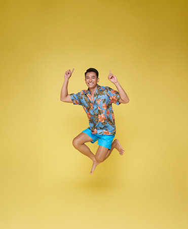 Full body young asian tourist man wear beach shirt jumping and pointing finger up isolated on yellow...