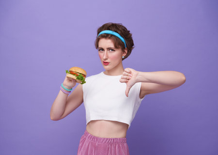 Young sad woman hold eat burger show hand stop gesture say no isolated on purple background. Proper...