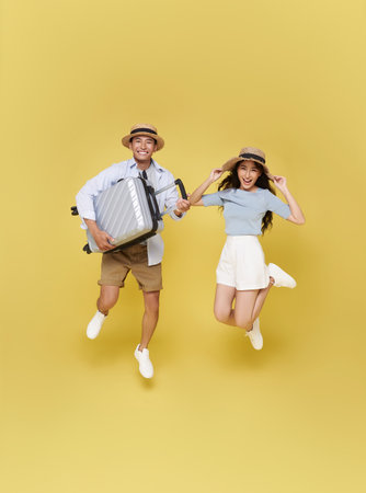 Happy fun Asian couple traveling vacation portrait jumping with suitcase ready for travel isolated on yellow studio background.の写真素材