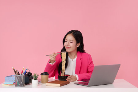 Beautiful young asian businesswoman eating noodles while work in office isolated on pink background.