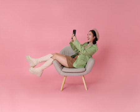 happy Asian woman wearing a green blazer, beret, and boots relaxing in a chair and looking at her phone isolated on pink background.の写真素材