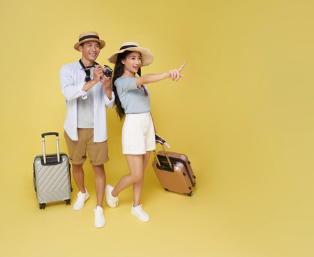 Happy Asian couple with hats luggage and camera pointing into distance Ready for adventure isolated on yellow background. Great for travel, holidays, or exploration themes.の写真素材