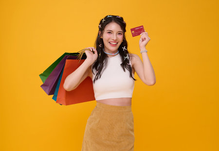 Happy Asian woman carrying colorful shopping bags and showing credit card shopping online isolated on yellow studio background.の写真素材