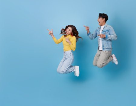 Happy Asian couple teenager jumping celebrating to travel on summer holiday and hand pointing finger isolated on blue copy space background.の写真素材