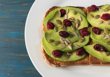 Vegetarian avocado toast with red cranberries, pumpkin seeds and sunflower seeds on a white plateの写真素材