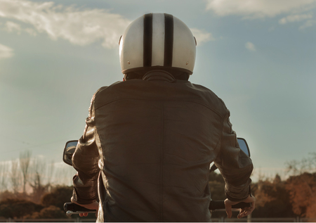 Man riding motorbike with brown leather jacket and helmet seen from behind, retro style