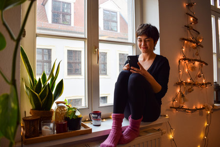 Young woman makes purchases in the Internet and pays by smartphone on Black Friday. Girl is enjoying buying gifts and sitting on comfortable windowsill at homeの写真素材