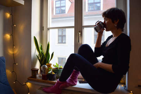 Meditative and hesitant girl drinks hot chocolate at the ledge next to some decoration while lost in imaginationの写真素材
