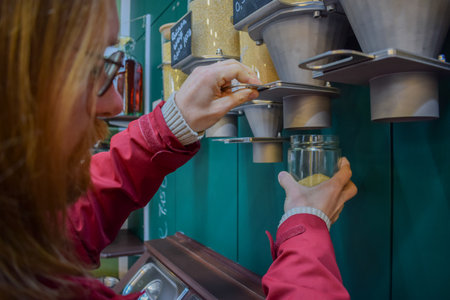Caucasian ginger client with glasses refilling a reusable jar with unpacked organic fresh groceries in a local marketの写真素材