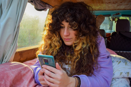 Independent curly haired woman text messaging mobile phone during a road trip sitting in resting in the bed of her vanの写真素材