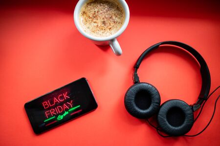 Mobile phone with Black Friday neon effect advertisement, headphones and cup of coffee on red background. Black Friday concept. Flat lay.の写真素材