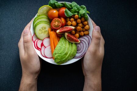 Caucasian man's hands holding a Buddha bowl with vegan ingredients on dark background.の写真素材