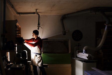 Portrait of working young male carpenter wearing protective clothes with a beard and a red sweater handles the wood on a lathe in the workshop. dark lighting, carpenters workshopの写真素材
