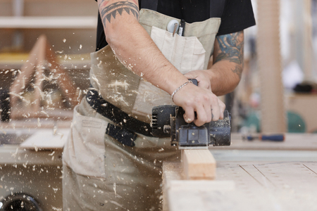 Carpenter working with electric planer on wooden plank in workshop. Hands and planer close up. sawdust flies from the working toolの写真素材
