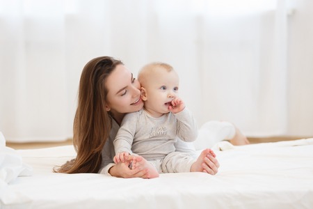Happy cheerful young mom smiling playing with her little baby boy lying in bed at home. family...