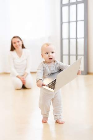 happy young mother and her little baby boy wearing white domestic clothes having fun with a laptop in the light living-room at homeの写真素材