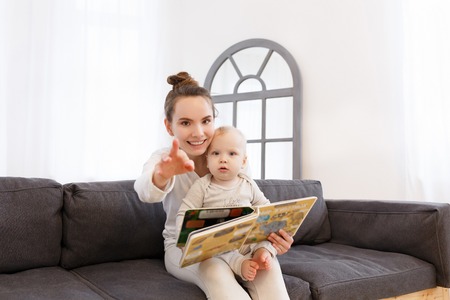 Pretty young mother reading a book to her little baby boy on a sofa in a light bedroom of house. Look at the camera and smile. Family portraitの写真素材