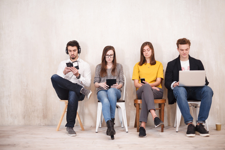 Four outsourcing employees sit together in creative studio and work with gadgets - smartphones,...