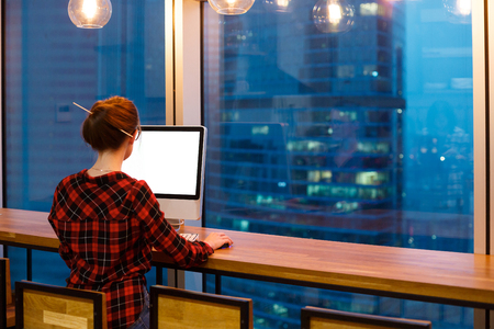 young unidentified hipster working behind large monitor, makes internet researches in office at night in the background of windows overlooking the big skyscrapersの写真素材
