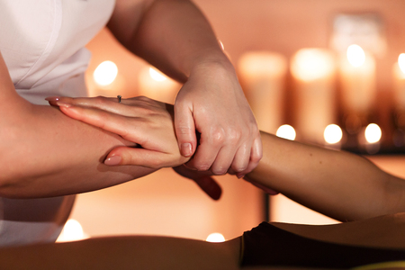 Hands of the masseur are doing massage on the hand of young unidentified woman lying on a massage table in spa salon against a blurred burning candle.Relax conceptの写真素材