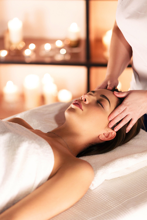 Female hands of masseuse doing massage on the head of beautiful young girl lying on massage table with closed eyes in a luxury spa resortの写真素材