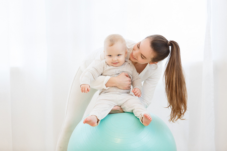 Happy young mother with baby wearing white sport clothes doing gymnastics with Exercise ball and having fun at light domestic home interior.の写真素材